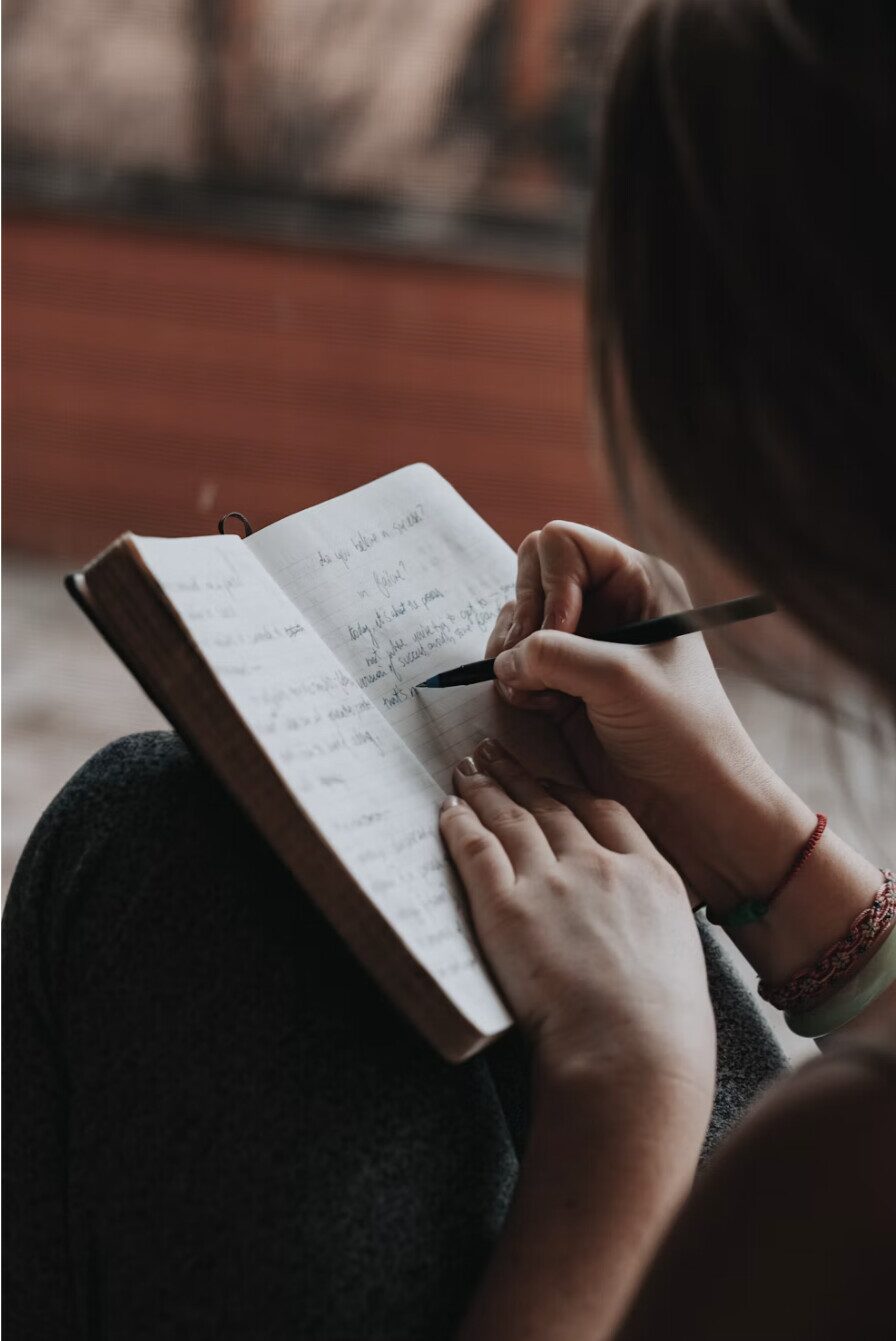An over-the-shoulder view of someone writing in an open journal, pen in hand, mid-reflection.